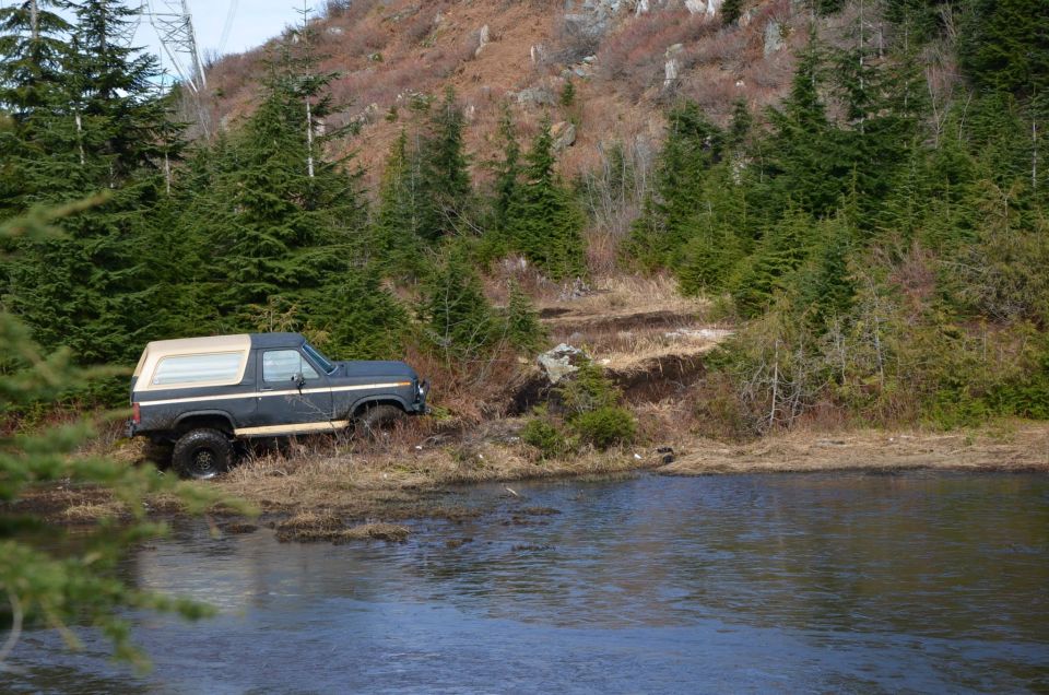 Dark blue Ford Bronco parked on a dirt path next to a body of water with trees and a hill in the background