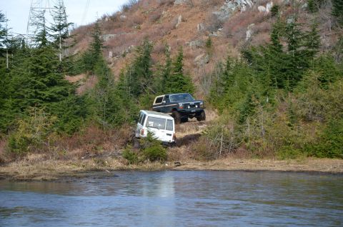 1984 Ford Bronco - Two SUVs navigate a steep, wooded incline next to a body of water.