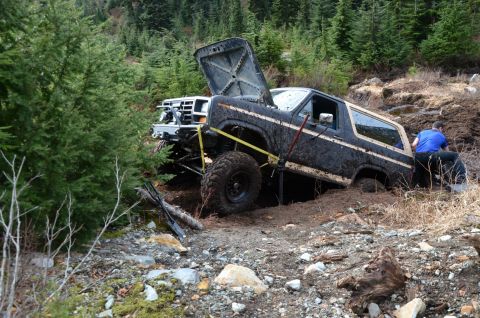 1984 Ford Bronco - A mud-splattered 1984 Ford Bronco is stuck in a muddy ditch with its hood open, while a person works to free it.