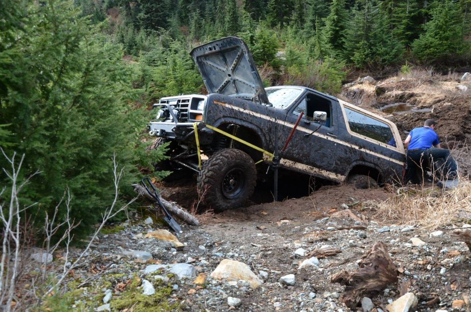 Mud-covered 1984 Ford Bronco stuck in a ditch, hood open, with a person attempting to free it