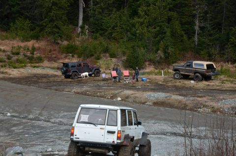 1984 Ford Bronco - Several off-road vehicles are parked in a clearing with people and camping chairs.