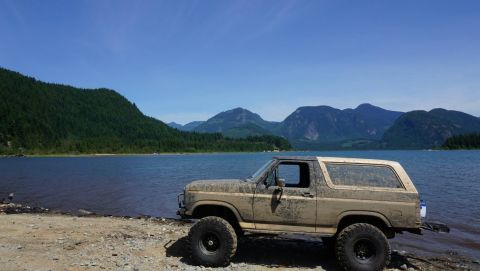 1984 Ford Bronco - A , mud-covered SUV sits by the water's edge, with a scenic mountain backdrop under a clear blue sky.