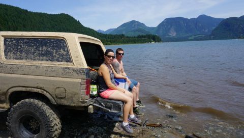 1984 Ford Bronco - Two people relax by a lake next to a dirty SUV. Mountains are visible in the distance.