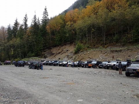 1984 Ford Bronco - A group of off-road vehicles are parked in a lot during autumn with colorful trees on the hillside.
