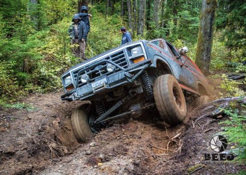 1984 Ford Bronco - A modified 1980s Ford Bronco navigates a challenging off-road obstacle in a wooded area.