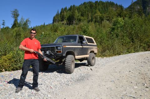 1984 Ford Bronco - A man stands with a shotgun next to a lifted Ford Bronco on a dirt road with a wooded hillside in the background.