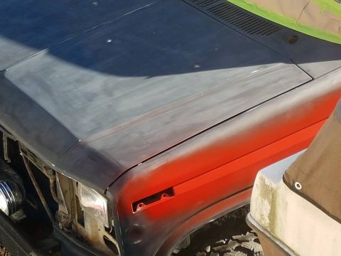1984 Ford Bronco - The hood and fender of a 1984 Ford Bronco show areas of gray primer and red primer, indicating preparation for paint.