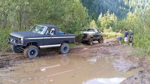 1984 Ford Bronco - Two off-road vehicles navigate a muddy trail with trees in the background.