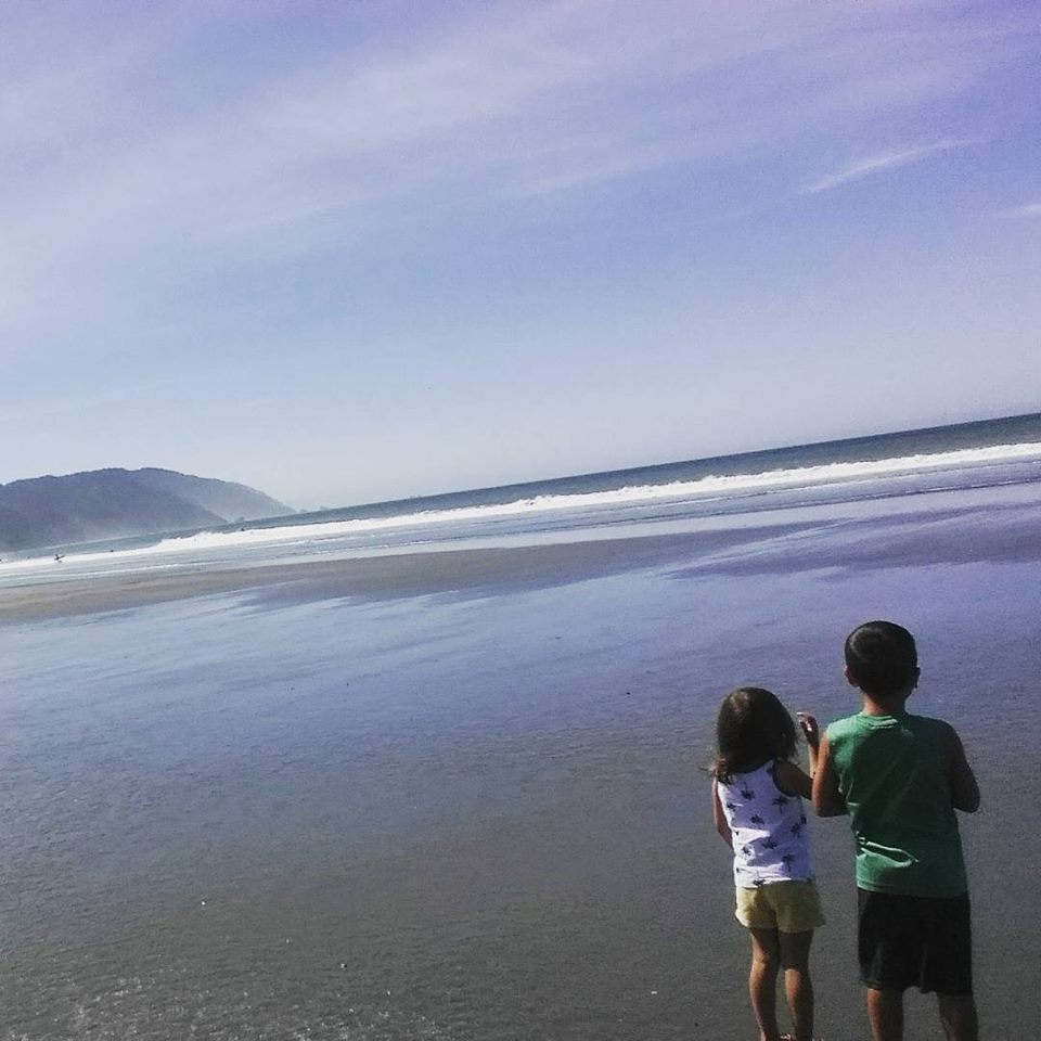 Two children stand on a wet sandy beach looking out at the ocean waves under a clear blue sky
