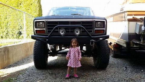1984 Ford Bronco - A young girl stands before a lifted Ford Bronco, dwarfed by its imposing front end and off-road tires.