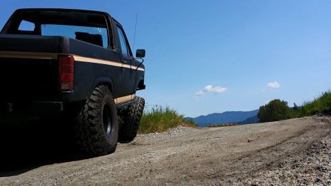 1984 Ford Bronco - Modified black Ford Bronco with tan stripe parked on a gravel road with mountains in the background.