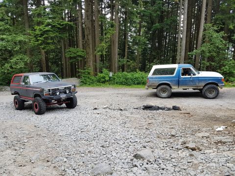 1984 Ford Bronco - Two modified off-road vehicles, a Ford Bronco and a Ford pickup, are parked on a gravel surface near a forest.