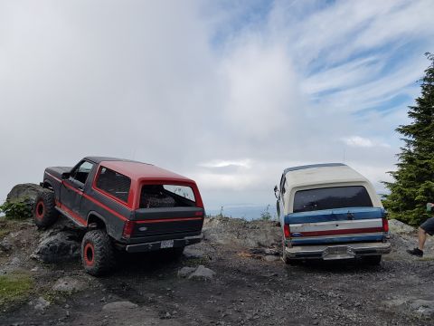 1984 Ford Bronco - Two off-road SUVs are parked on a rocky mountain path with a cloudy sky above.