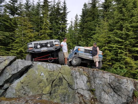 1984 Ford Bronco - Two men stand by their heavily modified trucks, parked on a rocky incline amidst a forest.