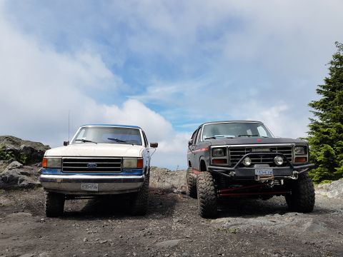 1984 Ford Bronco - Two SUVs, likely from the 1980s, are parked side-by-side on a rough, rocky terrain.