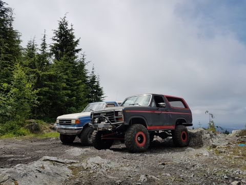 1984 Ford Bronco - Two modified off-road vehicles are parked on a rocky terrain with a forest and cloudy sky in the background.