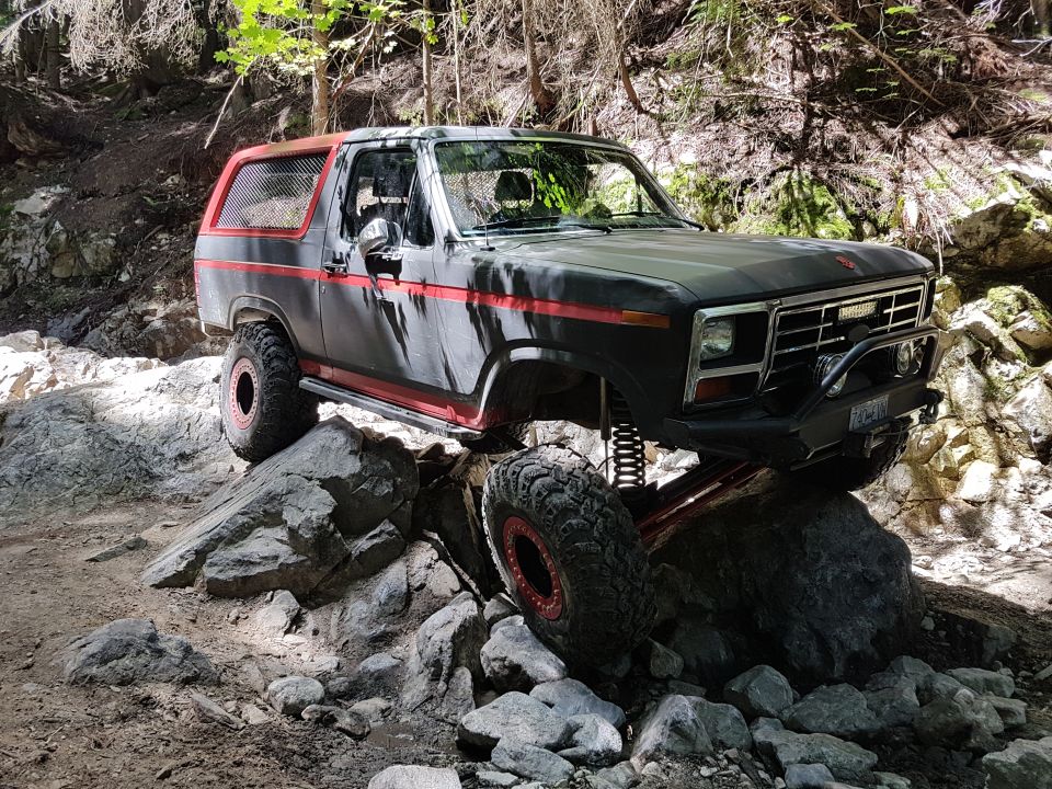 Modified Ford Bronco with red trim and off-road tires climbing over rocks on a trail