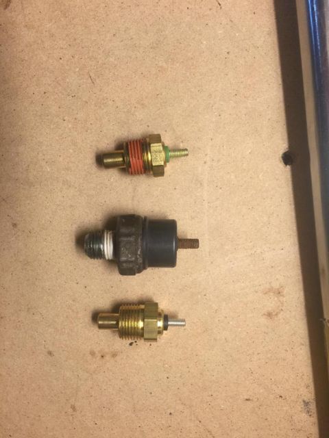 1989 Ford Bronco - Three threaded engine sensor switches on cardboard, including temperature-style senders with brass and black housings.