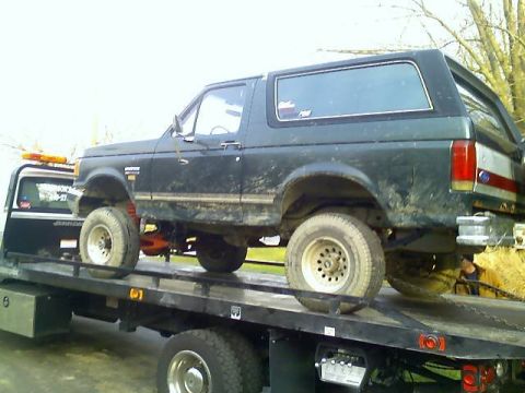 1989 Ford Bronco - Muddy green lifted Ford Bronco with a half cab top strapped onto a flatbed tow truck.