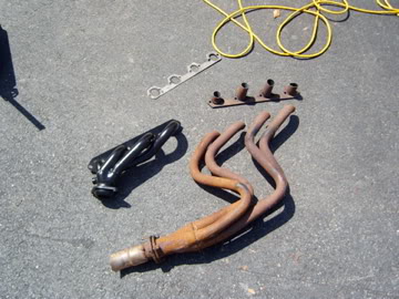 1989 Ford Bronco - Two exhaust manifolds, a gasket, and bolts are laid out on an asphalt surface, likely for a vehicle repair or upgrade.