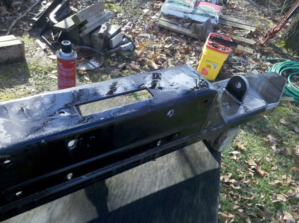 Black metal bumper with tow hitch attachment and rectangular cutout, resting on a wooden surface outdoors