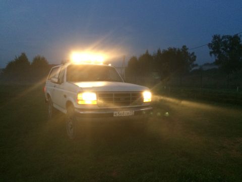 1993 Ford Bronco - A white 1993 Ford Bronco is parked in a grassy field at dusk, its light bar and headlights illuminated.