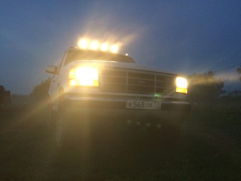 1993 Ford Bronco - White Ford truck with a light bar on the roof, seen at dusk with its headlights illuminated.
