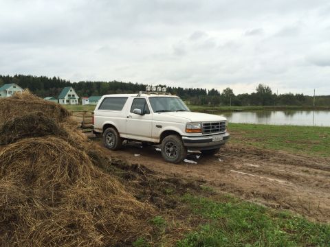 1993 Ford Bronco - A white Ford Bronco is parked on a dirt path next to a large haystack, with a lake and houses in the background.