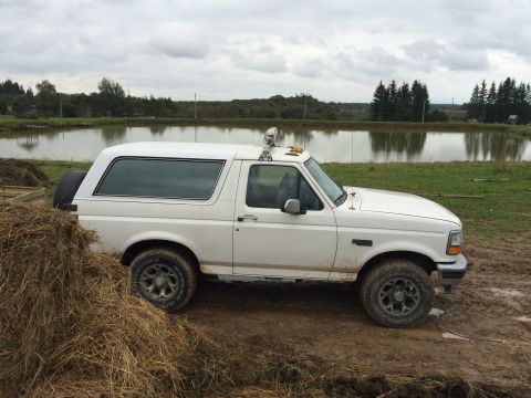 1993 Ford Bronco - A white 1993 Ford Bronco is seen parked on muddy terrain beside a calm body of water under an overcast sky.