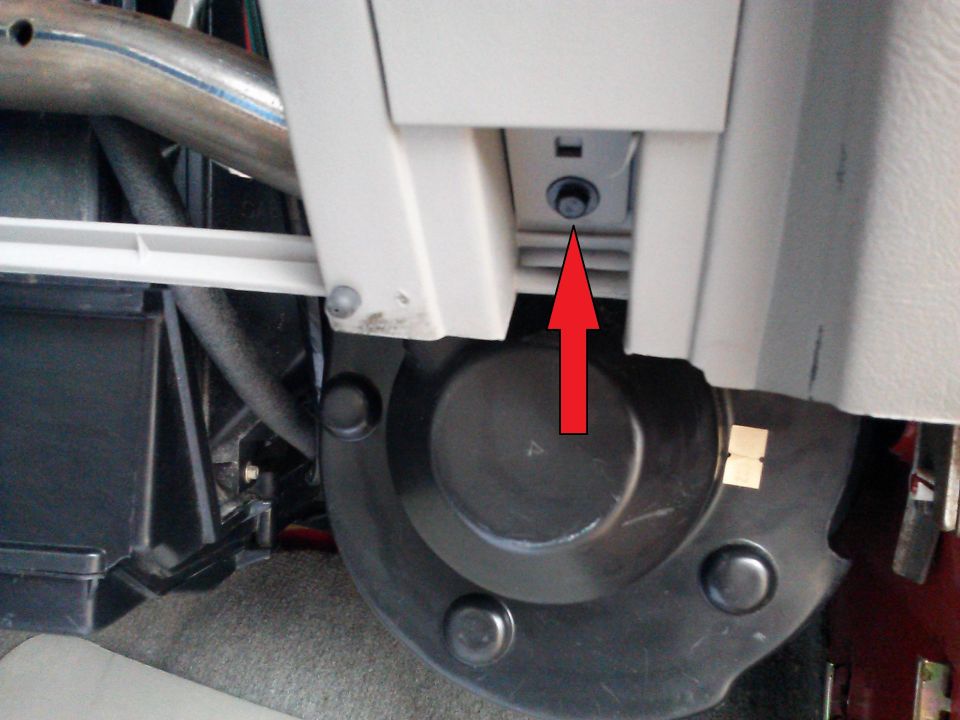 Interior view of beige dashboard panel with a red arrow pointing to a bolt near a black circular fan housing