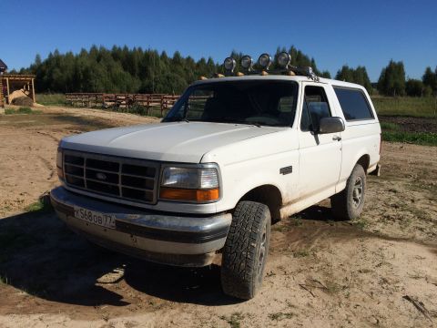 1993 Ford Bronco - A white 1990s Ford Bronco is parked on a dirt road with a wooded area and a fence in the background. The vehicle has a roof-mounted light b…