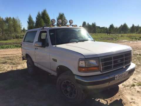 1993 Ford Bronco - A white 1993 Ford Bronco is parked on a dirt road in a grassy field with trees in the background. The vehicle has a roof rack with four aux…