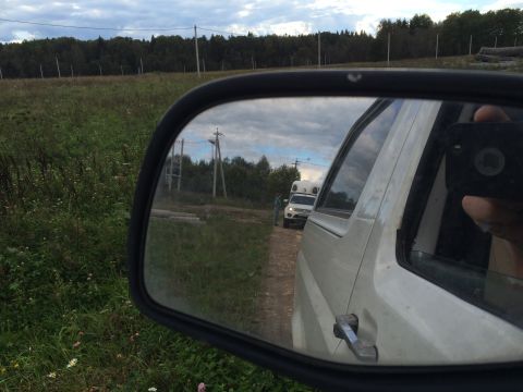 1993 Ford Bronco - A side mirror reflects a white SUV with a camper shell on a dirt road, with a person standing nearby. The reflection also shows a wooded ar…