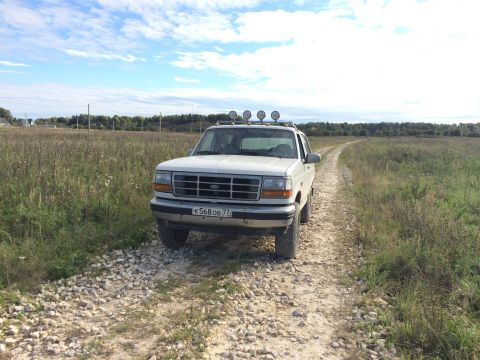 1993 Ford Bronco - A white 1993 Ford Bronco is parked on a gravel road in a grassy field under a partly cloudy sky. The vehicle has four auxiliary lights moun…