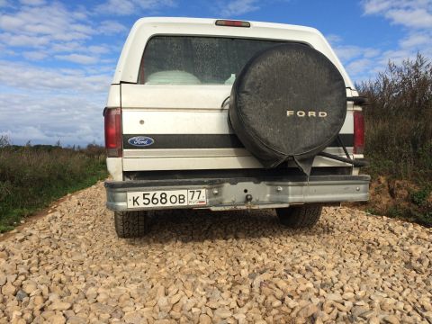 1993 Ford Bronco - The rear view of a white Ford Bronco is shown parked on a gravel road with a spare tire mounted on the back. The vehicle has a Russian lice…
