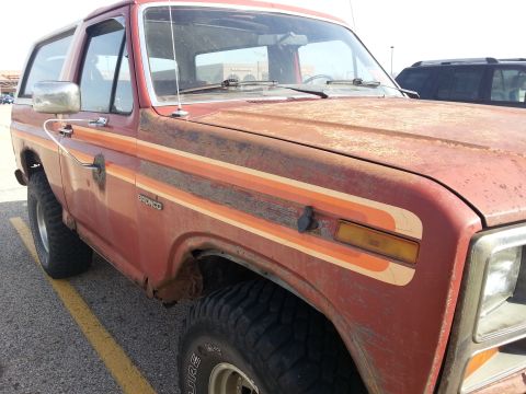 1984 Ford Bronco - Photograph of a weathered red Ford Bronco showing the front fender, door, and side stripe, not a document.