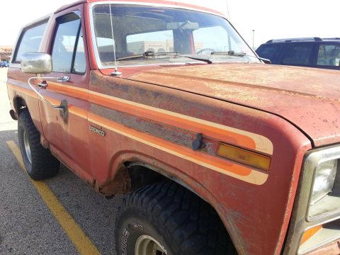 1984 Ford Bronco - Photograph of a weathered red Ford Bronco showing the front fender, door, and side stripe, not a document.
