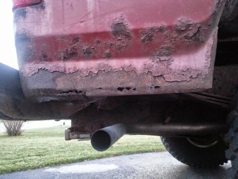 1984 Ford Bronco - Photograph of rust damage on a red vehicle's lower body near the exhaust, not a document.
