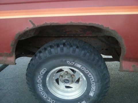 1984 Ford Bronco - Photograph of a rusted rear wheel well and tire on a red vehicle, not a document.