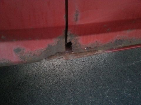 1984 Ford Bronco - Photograph of rust-through damage at the base of a red body panel, not a document.