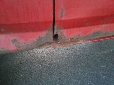1984 Ford Bronco - Photograph of rust-through damage at the base of a red body panel, not a document.