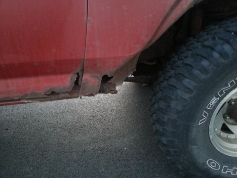 1984 Ford Bronco - Photograph of rust-through holes in a red rocker panel near the rear wheel, not a document.