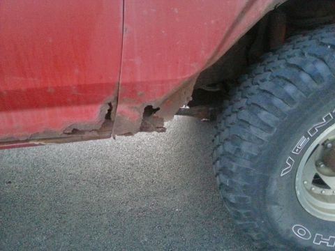 1984 Ford Bronco - Photograph of rust-through holes in a red rocker panel near the rear wheel, not a document.