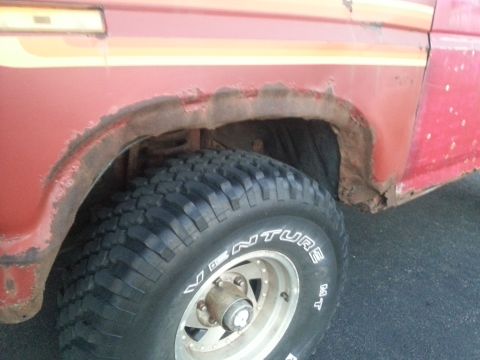 1984 Ford Bronco - Photograph of a rusted front wheel well and tire on a red vehicle, not a document.