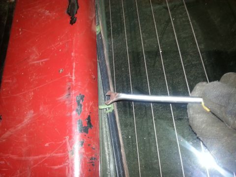 1984 Ford Bronco - Photo of a hand prying along the rear glass channel and weatherstrip of a red vehicle, not a document.
