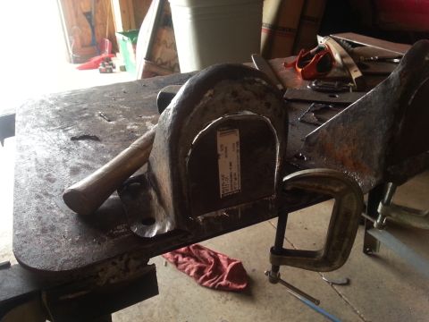 1984 Ford Bronco - Metal part with a label and a hammer on a workbench, clamped in place for work.
