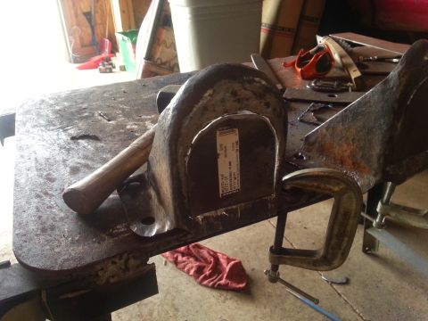 1984 Ford Bronco - A metal bracket is secured in a vise on a workbench, with a hammer resting on the surface. A label is attached to the bracket.