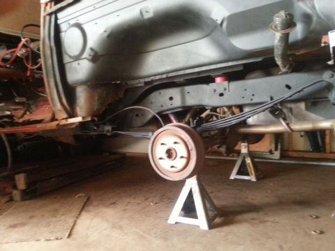 1984 Ford Bronco - The undercarriage of a vehicle is shown, with a rear drum brake assembly supported by a jack stand and leaf springs visible.