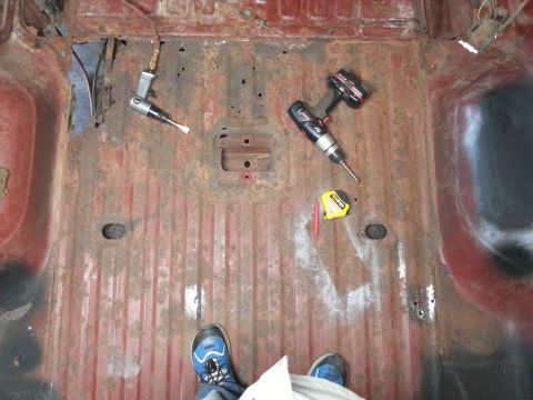1984 Ford Bronco - Workshop photo of a rusty cab floor pan with a drill and tape measure, not a technical document.
