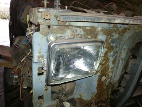 Square sealed-beam headlight mounted in a rusty front fender support with wiring